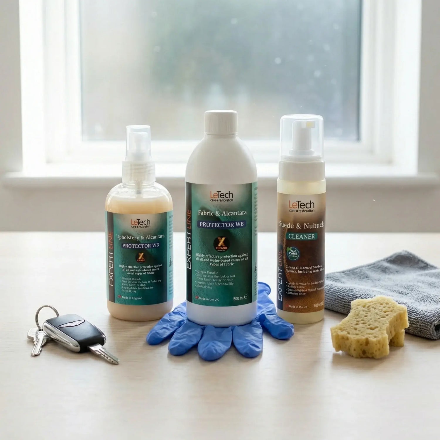 Three bottles of LeTech Leather cleaning products on a surface with gloves, keys, and a sponge in front of a window.