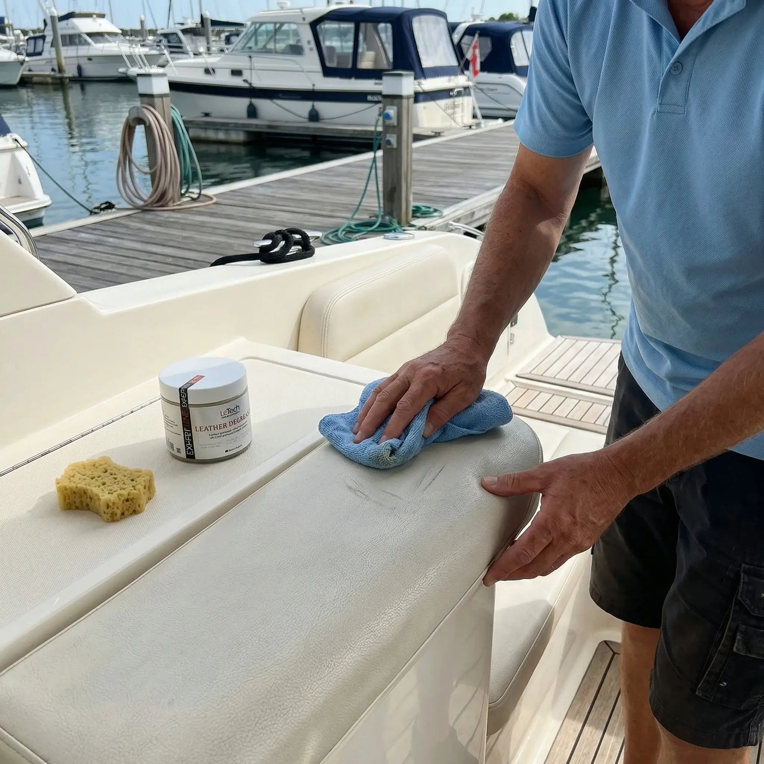 Person cleaning a boat's white surface with a cloth and a container of cleaning product, docked boats in the background. Boat leather repairs by LeTech